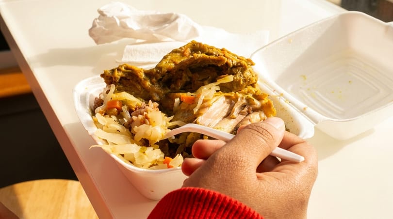 A person's hand holds a fork, eating curry chicken and rice from a white takeout container.