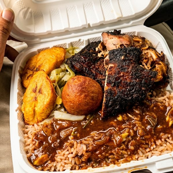 A hand with a smiley face tattoo holds a takeout container of jerk chicken, rice, plantains, and a dumpling.